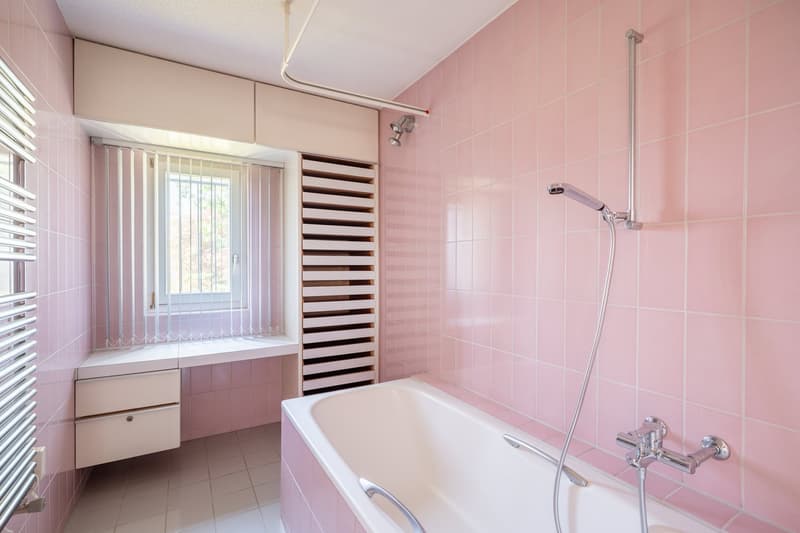 Bathroom with pink tiles, white bathtub, shower head, white sink, window with blinds, towel rack