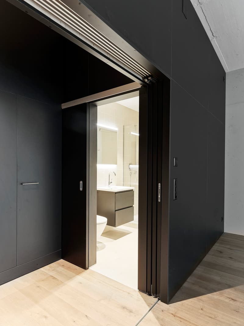 Modern bathroom with white tiles, large mirror, vanity with sink and drawers, toilet, open sliding door to hallway, wooden flooring.