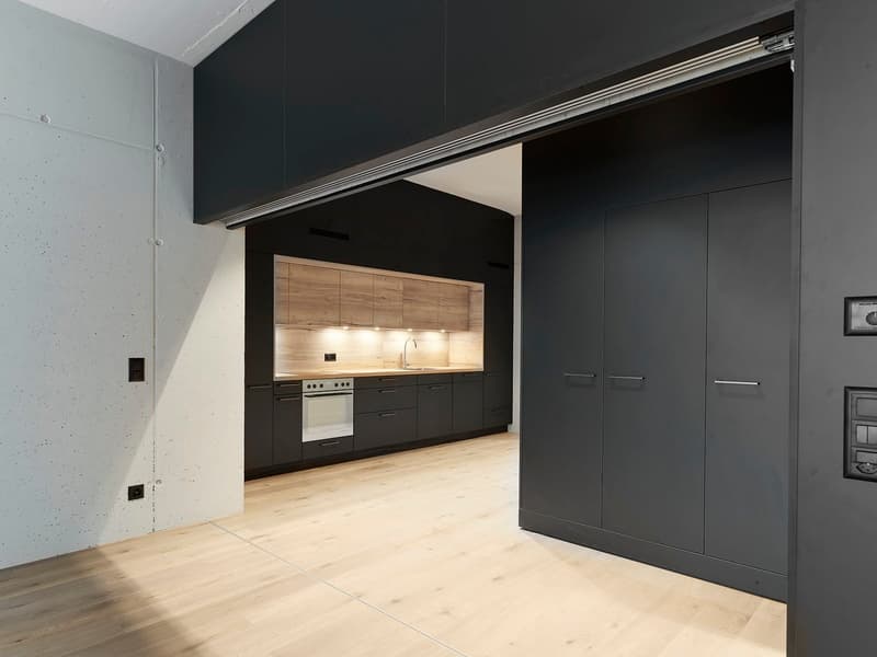 Modern kitchen with black cabinets, wooden countertops, and built-in appliances including a dishwasher. The kitchen is open to a living space with light hardwood flooring.