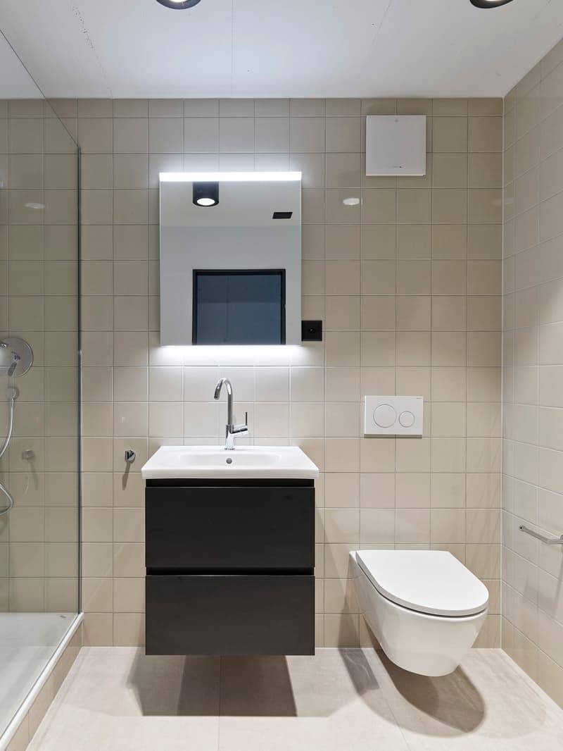 Modern bathroom with white tiles, black vanity unit with sink, wall-mounted toilet, and recessed lighting above the mirror.
