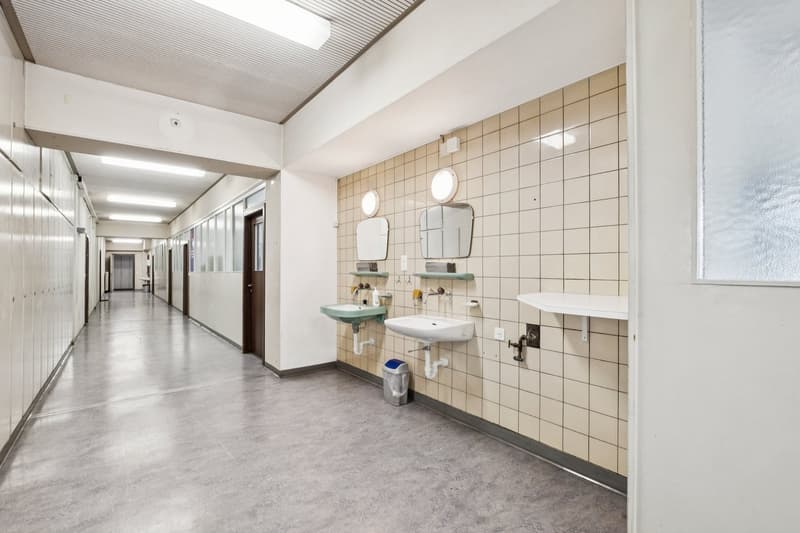 Hallway with tiled walls, ceiling lights, row of lockers, two sinks with mirrors and shelves, trash cans, and a window.