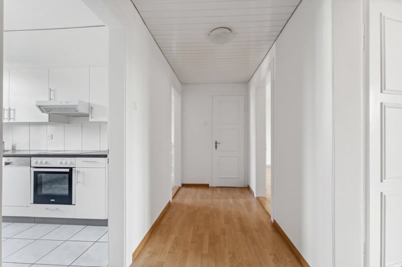 Empty hallway with wooden floor, white walls, kitchen with oven and dishwasher, white cabinets, white tiled floor in kitchen