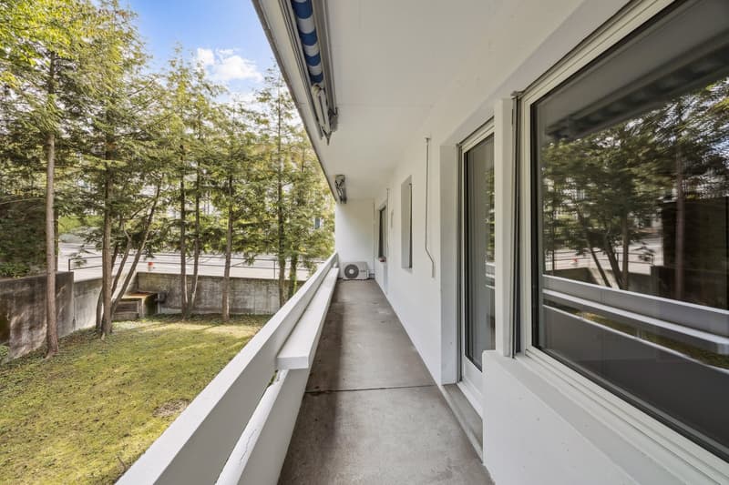 Balcony with white railing, concrete floor, large windows, air conditioner, trees, grass