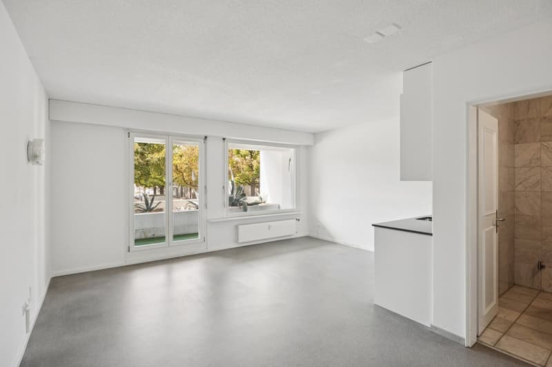 Empty room with white walls, gray floor, large windows with sliding doors leading to a balcony, and a white counter with a sink.
