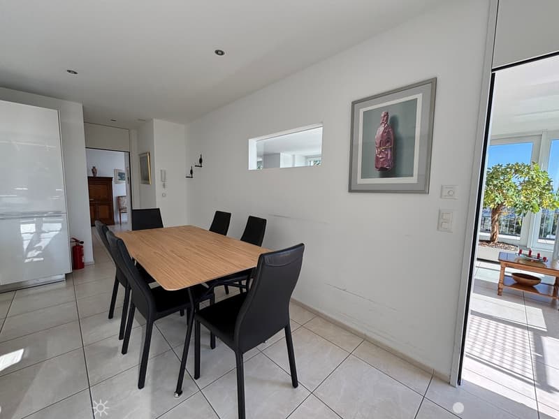 A dining room with a wooden table, black chairs, white walls, and tiled floor, leading to a balcony with a view and a wooden table with a bowl.