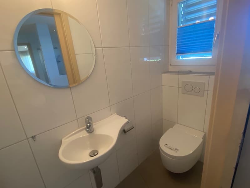 Small bathroom with white tiled walls, white sink, round mirror, toilet, blue window blinds, and wooden frame around the mirror.
