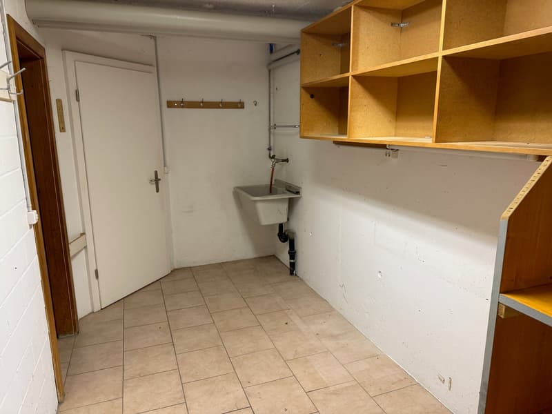 Small, tile floored, white-walled room with a door, wall mounted shelves, a sink with drain, a pipe, a wall mounted clothes rack, and a power outlet