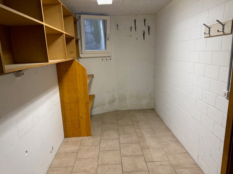 Empty room with tiled floor, white brick walls, wooden shelving, small window, ceiling light, hooks mounted on the wall.