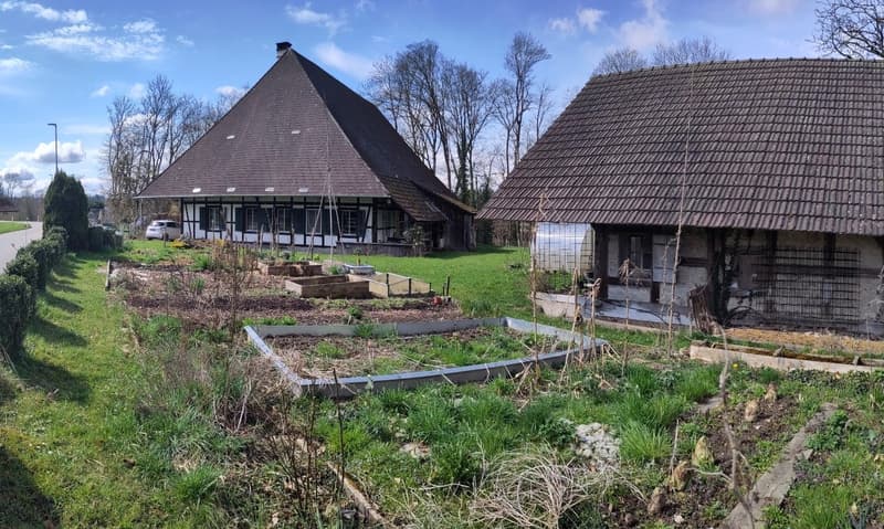 Two-story farmhouse with a steep, tiled roof, surrounded by trees and a garden area. The house has a raised ground floor and a parking space nearby. The exterior features a traditional half-timbered design.