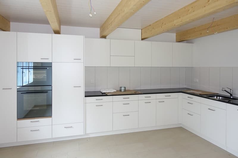 Modern kitchen with white cabinetry, black countertops, stainless steel sink, double oven, dishwasher, wooden beams