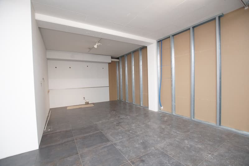 Empty room with grey tiled floor, white walls, and metal structures. Wires visible in corners.