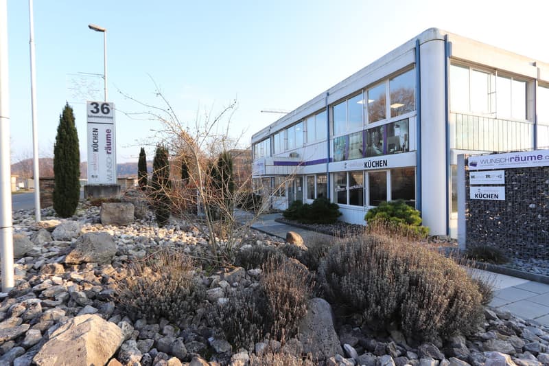 Modern building with glass windows, sign indicating 'exklusiv Küchen', surrounded by rocks and plants