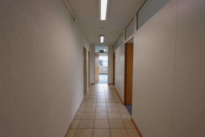 Empty hallway with white walls, tiled floor, ceiling lamps, open doors at both ends.
