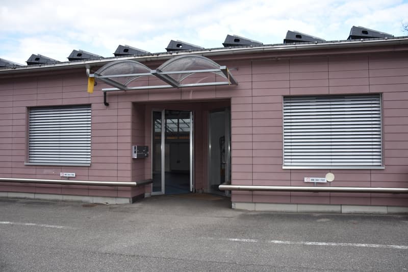 Pink building with solar panels on the roof, glass entrance, metal railings, and two windows with blinds.