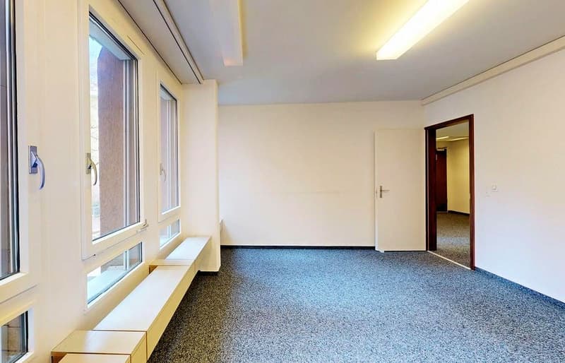 Empty room with white walls, ceiling, and windows. Carpeted floor. Benches along the wall. Two doors.
