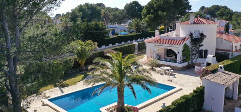Two-story Mediterranean-style villa with red tile roof, surrounded by lush greenery and palm trees. The property features a large swimming pool and a balcony or terrace overlooking the pool and garden area.