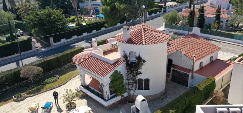 Two-story Mediterranean-style villa with red tile roof, white stucco walls, and arched windows. The house has a balcony or terrace, a garage, and a parking area. The property is surrounded by lush greenery and landscaping.