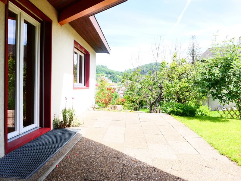 Two-story wooden house with red trim, surrounded by lush greenery and a paved patio area. The house is situated on a raised ground floor, providing a scenic view of the surrounding landscape.