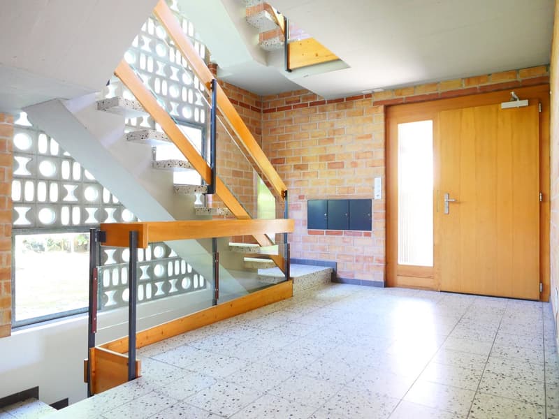 The image shows the interior of a building with a unique architectural design. The space features exposed brick walls, a wooden staircase with a metal railing, and a tiled floor. There are several storage units or lockers visible on the wall. The room app