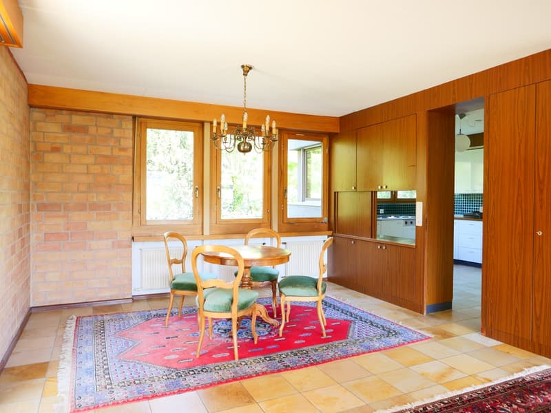 Spacious dining area with wooden paneled walls, large windows, a chandelier, a round wooden dining table with chairs, and a colorful patterned rug on the tile floor. The kitchen can be seen through the doorway.