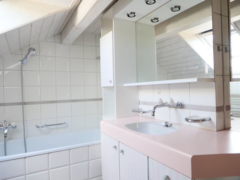 The image shows a bathroom with white tiled walls, a vanity sink with a pink countertop, and a shower area with a glass door. The bathroom has recessed lighting fixtures on the ceiling and a mirror above the vanity sink.