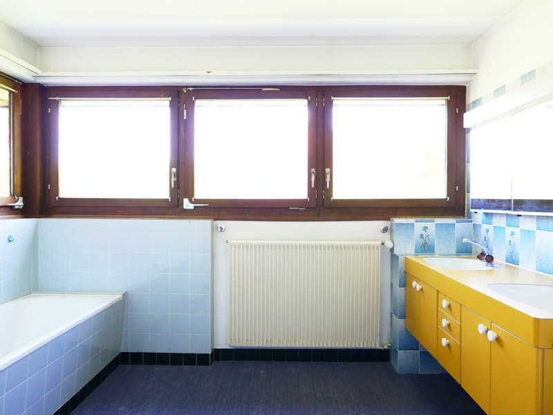 Spacious bathroom with large windows, white tiles, yellow vanity cabinet, and a radiator
