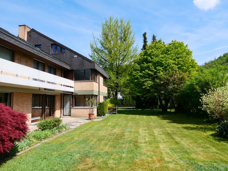 2-story house with a slanted roof, brick and wood exterior, large windows, surrounded by a well-maintained lawn and lush greenery including trees and shrubs. The property appears to have a balcony or terrace and offers a scenic view of the surrounding nat