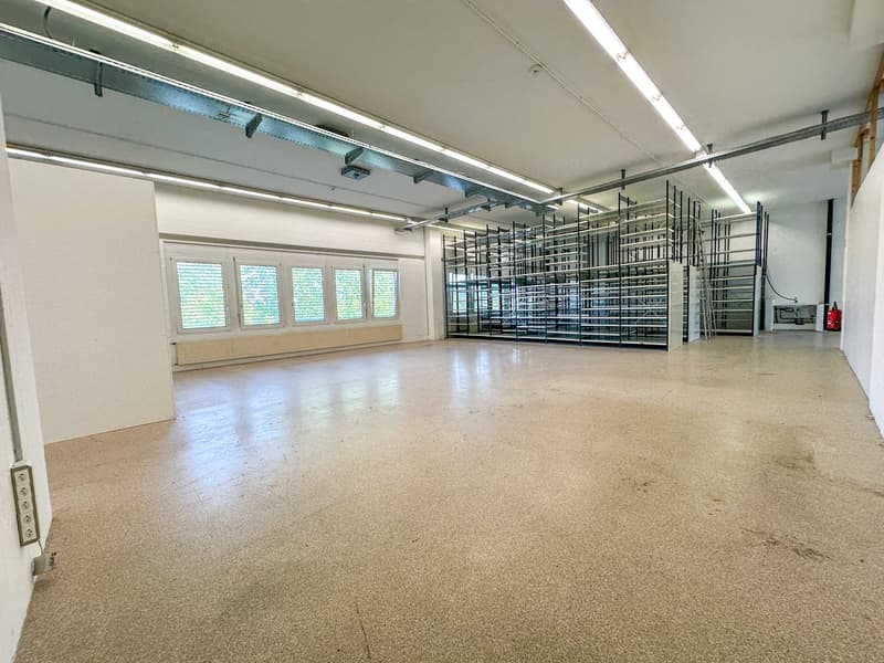 Large open commercial space with concrete flooring, metal shelving units, and windows along one wall. The space appears to be a warehouse or storage facility.