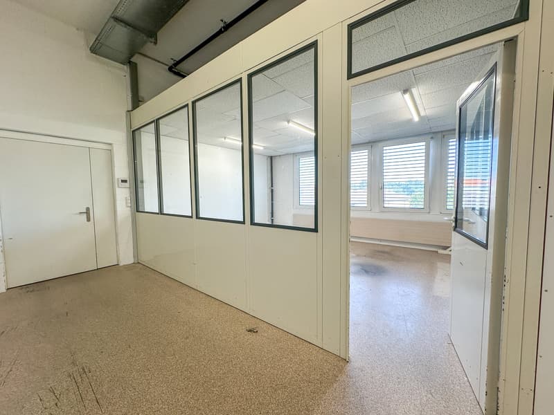 The image shows an empty office space with white walls, a glass partition, and a door leading to another room. The space has a concrete floor and fluorescent lighting fixtures on the ceiling.