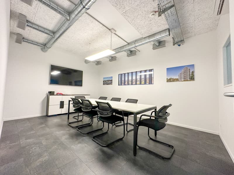 empty room, rectangular table, black office chairs, white walls, flat screen television mounted on wall, ceiling mounted lamps, light wooden desk on left
