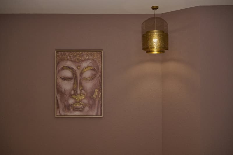A beige corner wall with a golden framed artwork of a Buddha and a golden hanging lamp.