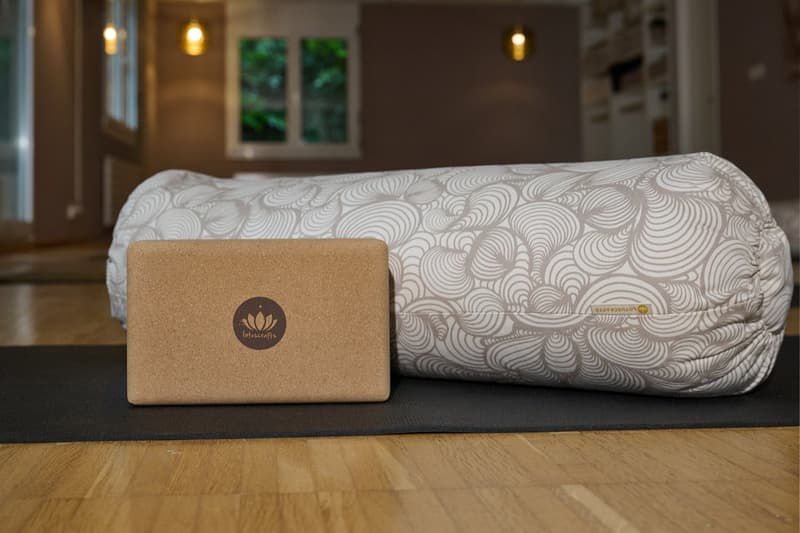 Yoga mats on wooden floor, brown yoga block, white yoga bolster, large window, shelves, radiator