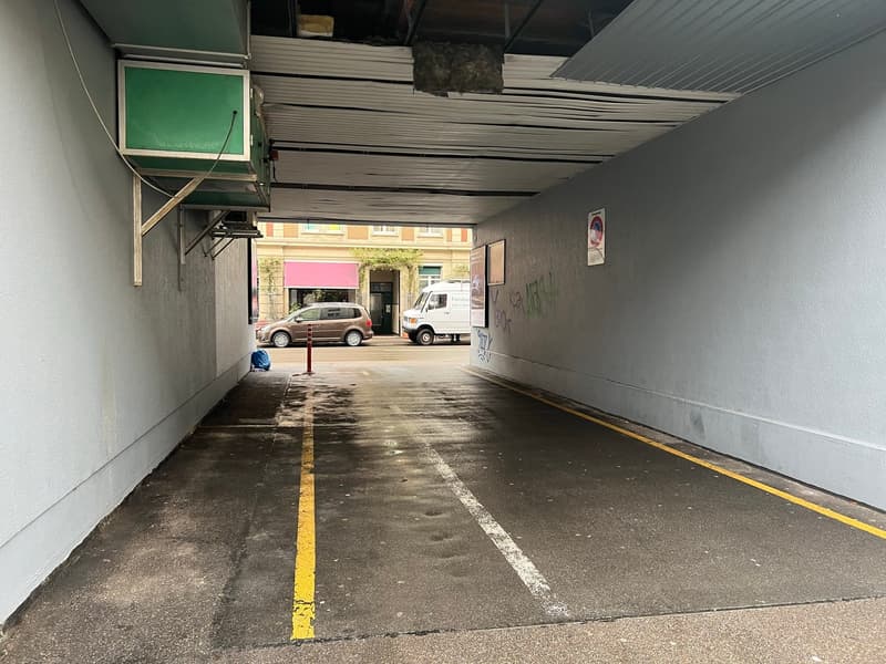 Parking space with concrete walls, ceiling with ducts, yellow and white lines on the floor, graffiti on the wall, and vehicles parked outside.
