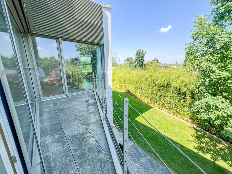 Balcony with glass walls, metal railings, tiled floor, green lawn, hedges, trees, clear sky