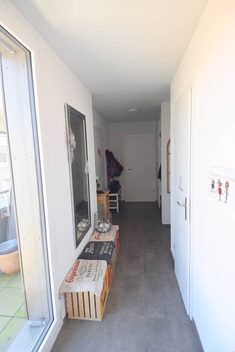 A hallway with white walls, tiled flooring, a mirror, storage baskets, a bench, and a glass door.
