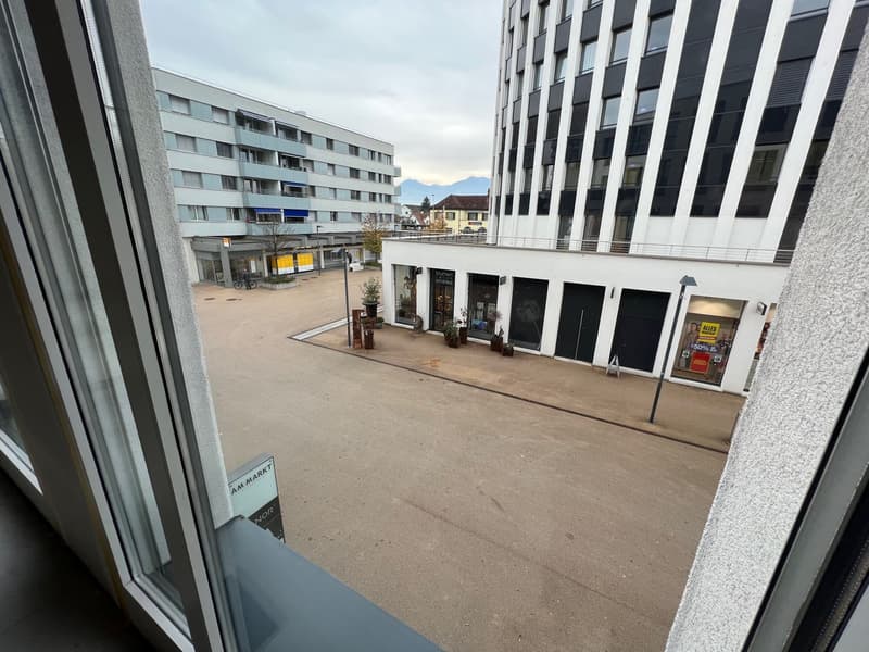 A street view from a window with a modern building, parking area, and mountains in the distance