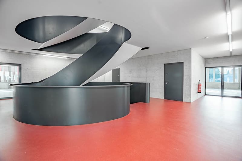 Open space with a large spiral staircase, concrete walls, red floor, two doors, fire extinguisher, windows