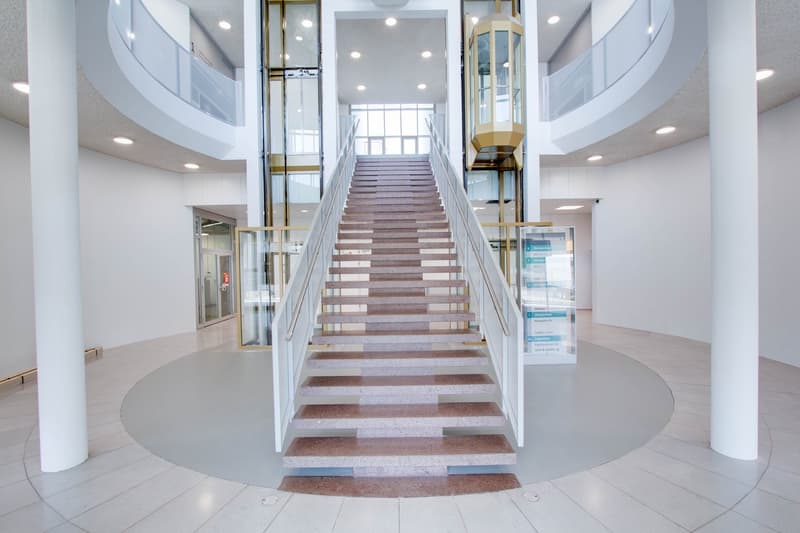 Large open area with marble steps and stainless steel railings, elevator on the right, glass walls and ceilings, white walls, circular tiles at the base of the steps.