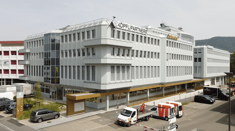 multi-story office building, white exterior, glass windows, red sign reading 'KOPFLI PARTNERS', multiple parked vehicles in the front