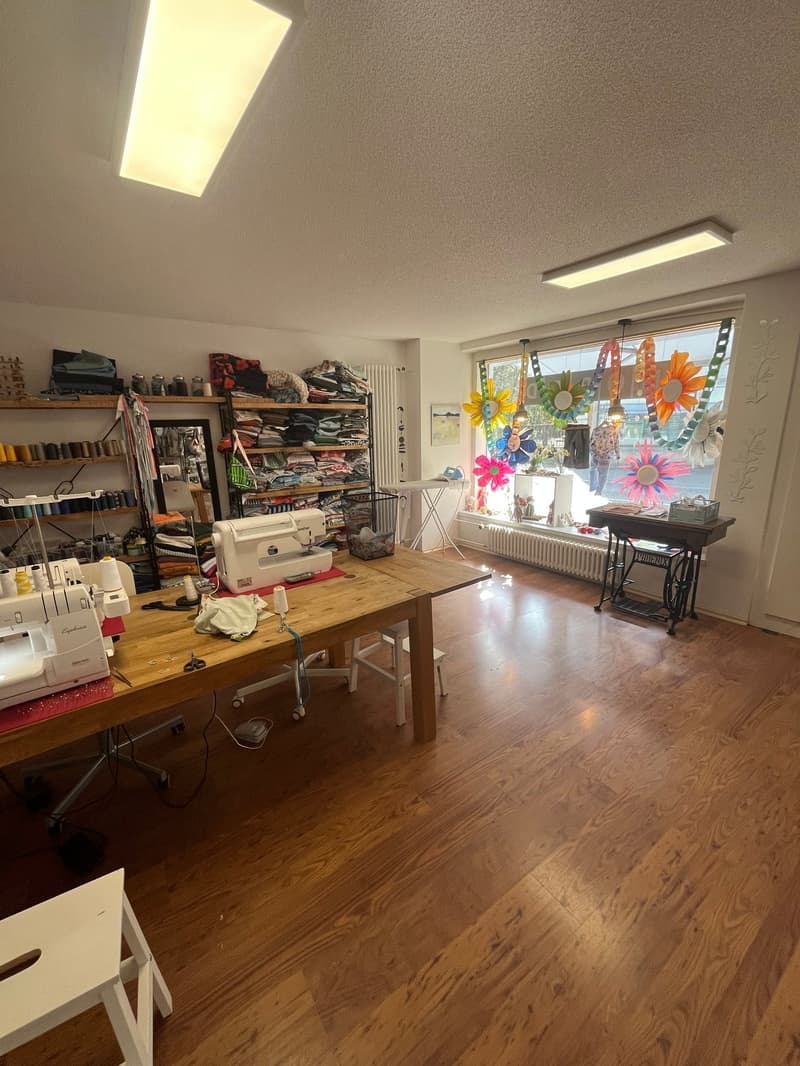 A room with a wooden table, sewing machines, shelves with clothes, a mirror, and a window with decorations.