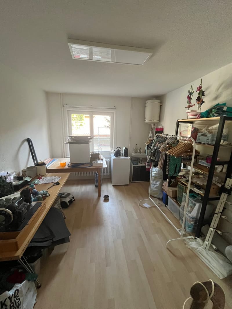 Empty room with wooden floor, white walls, two tables, clothes rack, shelves, radiator, window, white cabinet, sink, teapot, storage boxes, and various items.