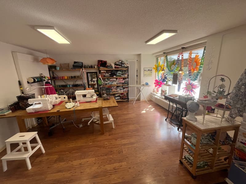 A room with a wooden floor, white walls, and a large window with colorful decorations. There are two tables with sewing machines, a shelf with various items, and a white stool. The room is well-lit with ceiling lights.