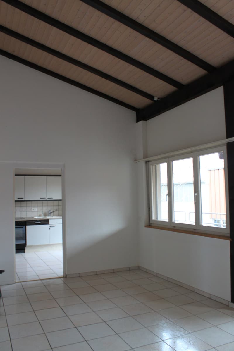 Empty room with white walls, tiled floor, and kitchen in the background. A window with a view outside.