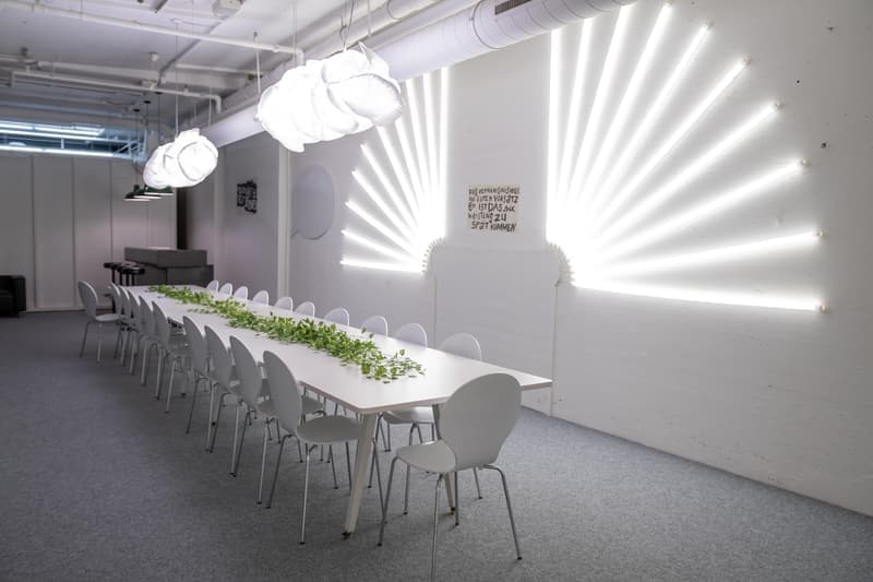Long dining table, white chairs, white lighting fixtures, white walls, green plants on table, abstract wall art, wooden floor.