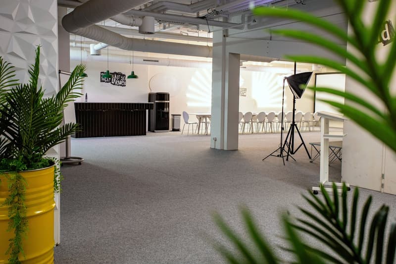 An empty office with white walls, carpet floor, tables and chairs, and some potted plants. In the background, there is a table with a white tablecloth and a black speaker. The room is equipped with modern equipment, such as a camera on a tripod and a blac