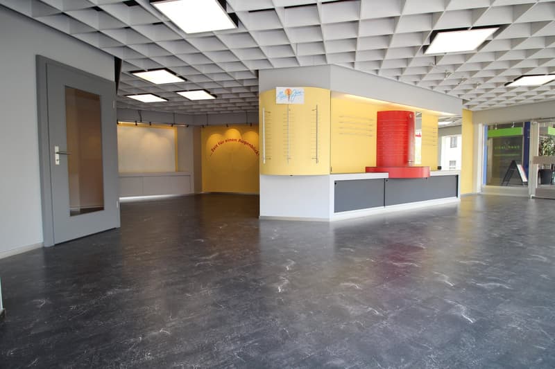 Large open office space with a grid-like ceiling, gray walls, and a reception desk with yellow and red accents. The floor is made of dark gray marble-like tiles. The space appears to be a modern and functional office environment.