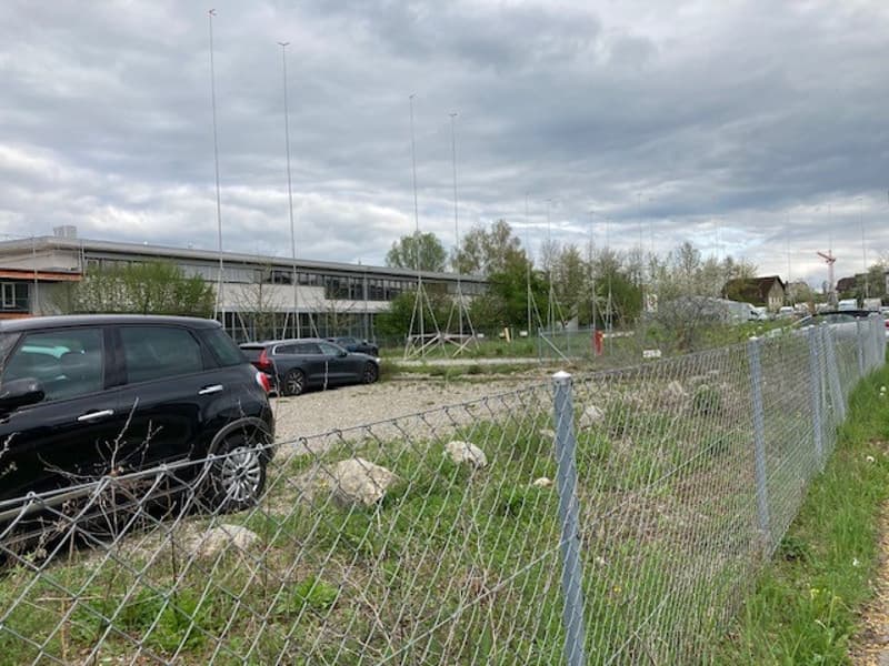 empty plot, chain link fence, parked cars, background is large building, cloudy sky