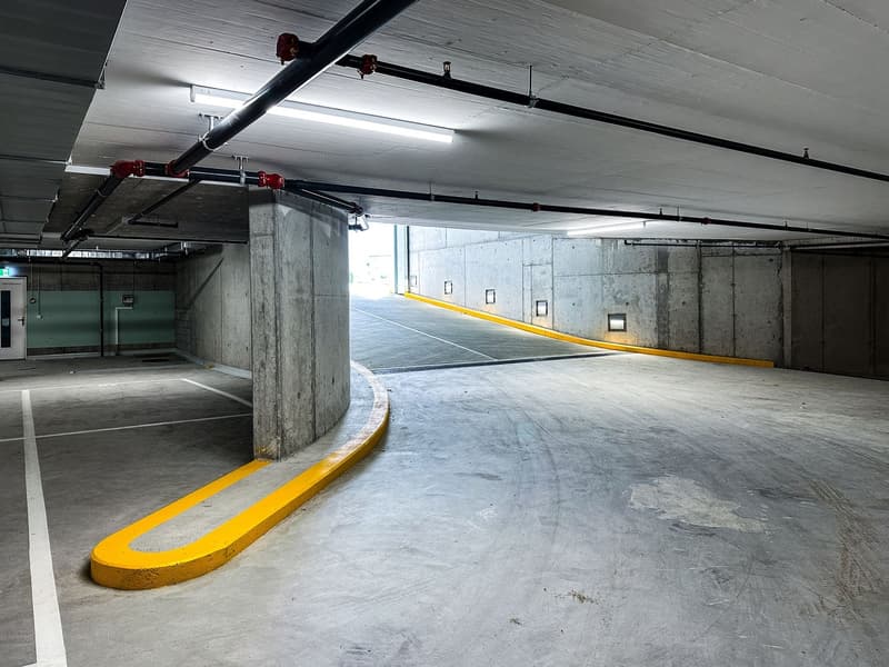 Empty parking lot, yellow and white curbs, concrete walls, ceiling with lighting and pipes, grey floor, exit door on left