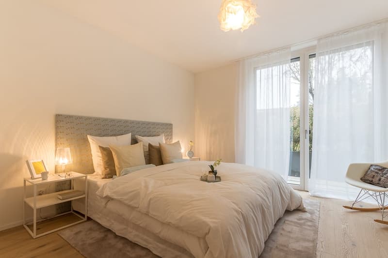 White walls, bed with pillows, nightstands with lamps, wooden floor, carpet, sliding glass doors leading to a balcony with railing