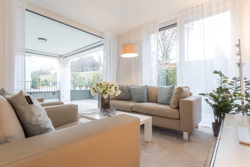 Beige leather couches, coffee table, floor lamp, balcony with metal railings, plants, white curtains, large windows, elevated floor